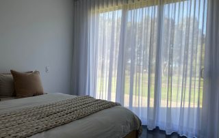 Bedroom with sheer curtains and cozy bedding. Sheer Curtain is supplied by C'Lect-a-Blind and made in Nettex Mykonos White