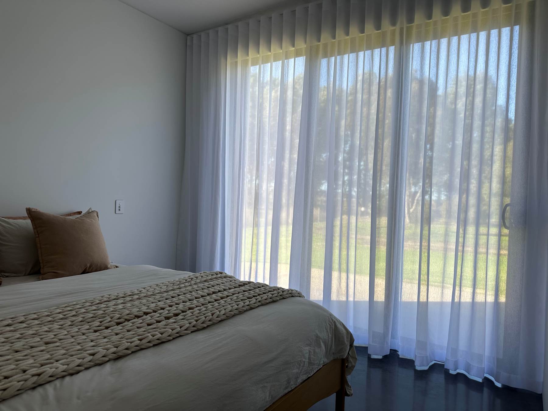 Bedroom with sheer curtains and cozy bedding. Sheer Curtain is supplied by C'Lect-a-Blind and made in Nettex Mykonos White